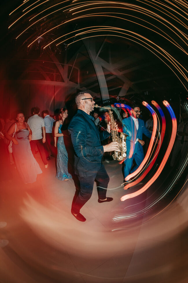 A saxophonist in a dark suit plays on a lively dance floor surrounded by people in formal attire, with swirling light trails creating a dynamic, festive atmosphere.