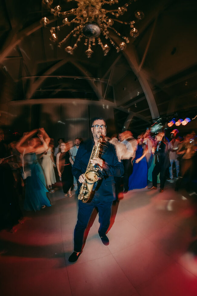 A man in a dark suit plays the saxophone at a lively party, with blurred dancers moving energetically around him under a chandelier in a modern, spacious venue.