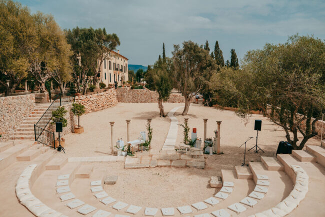 Outdoor amphitheater with stone seating arranged in a semicircle, a central aisle lined with white stones, surrounded by trees and stone buildings under a partly cloudy sky. Sound speakers and small decorations are set up.