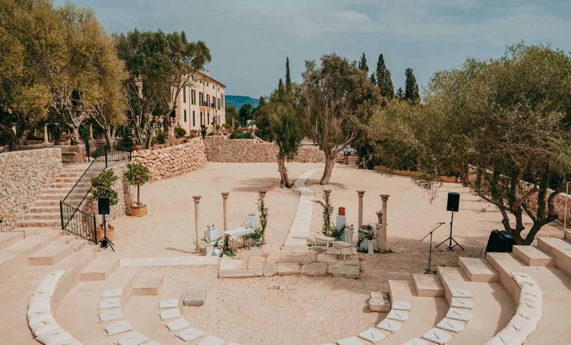Ein Amphitheater im Freien mit Sitzgelegenheiten aus Stein umgibt eine kreisförmige Sandfläche, die wunderschön mit Blumen und Säulen dekoriert ist - ein idealer Ort für eine Hochzeit auf Mallorca in ses cases de sa font seca, mit Olivenbäumen und Bergen als atemberaubender Kulisse.