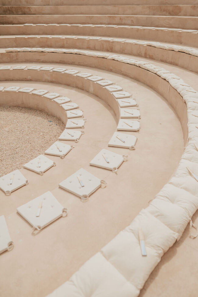 Cushioned stone steps in an amphitheater, each seat with a white notepad, wooden stick, and pen neatly placed on top, arranged in concentric circles.