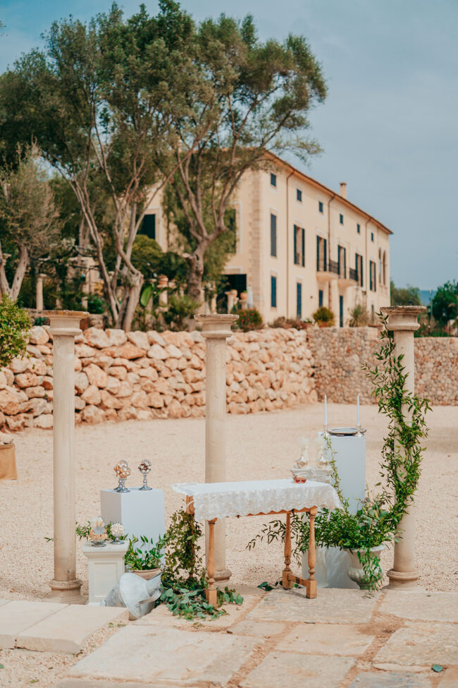 An outdoor wedding altar with a lace-covered table, candles, and greenery, set between two stone columns in front of a rustic stone building and a low stone wall, surrounded by trees.