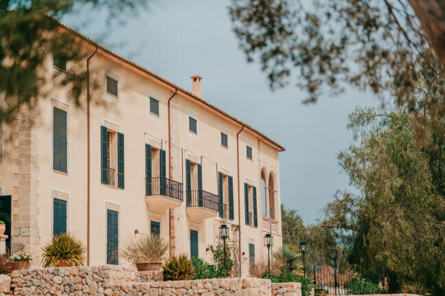 A large Mediterranean-style villa with light-colored walls, black shutters, small balconies, and arched windows, surrounded by trees and stone landscaping under a cloudy sky.