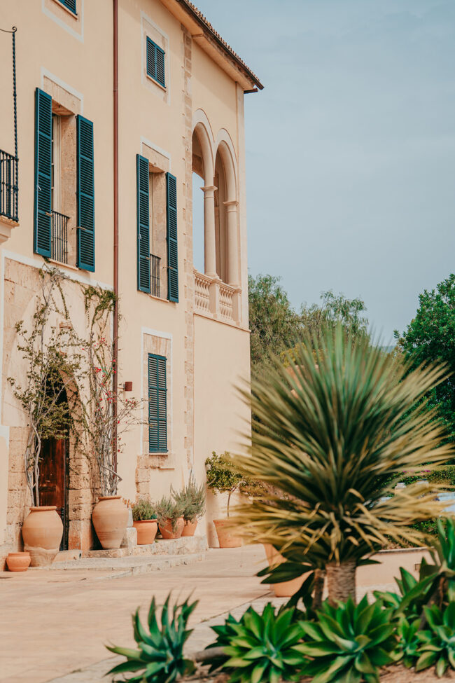 A Mediterranean-style building with tall windows and blue shutters, surrounded by terracotta pots with plants and greenery in the foreground under a partly cloudy sky.