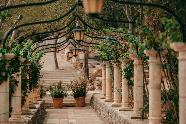 A stone walkway lined with columns and green vines, topped by black iron arches holding lanterns, leads through a garden with potted plants and trees in the background.