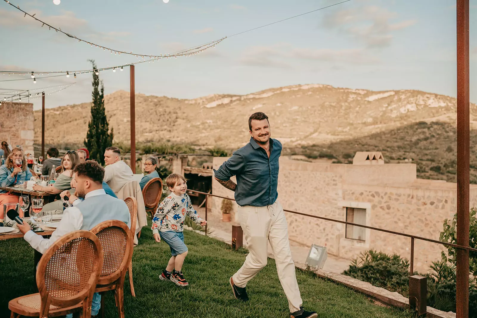 Ein lächelnder Mann in blauem Hemd und beiger Hose geht über das Gras, während ihm ein lachender Junge auf einer Ses Voltes Mallorca Hochzeit folgt, während die Leute an Tischen im Freien mit einer malerischen Berglandschaft im Hintergrund sitzen.