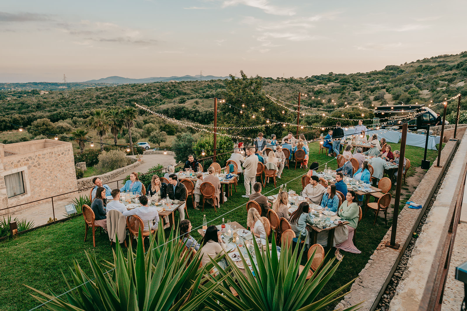 Ein Treffen im Freien bei Sonnenuntergang, bei dem die Gäste an runden Tischen auf einer grasbewachsenen Terrasse der Finca Ses Voltes Mallorca speisen, umgeben von üppigen Hügeln und Lichterketten über dem Kopf, die eine festliche und entspannte Atmosphäre schaffen.