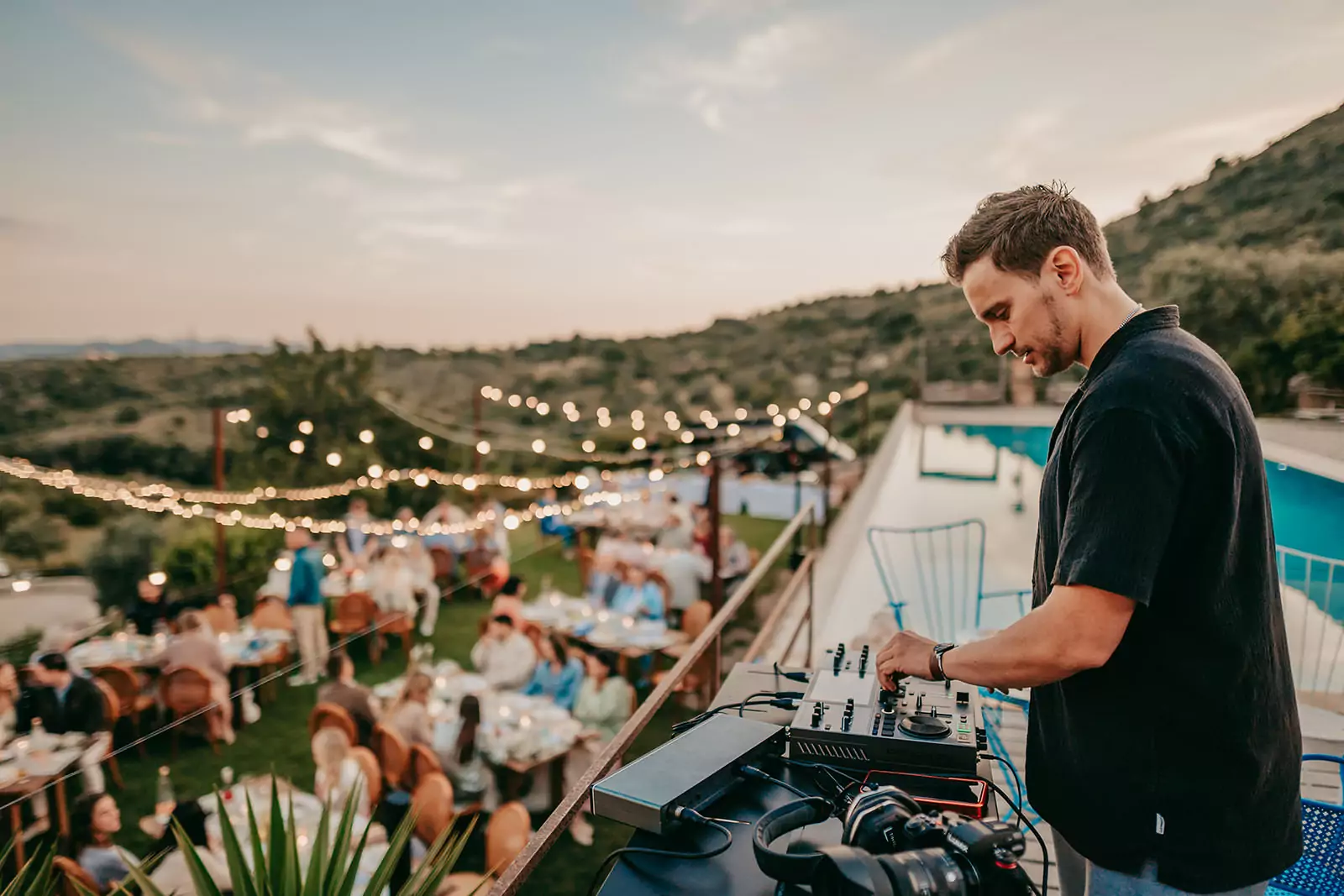 Ein DJ stellt die Regler eines Mischpults bei einer Veranstaltung im Freien auf der Finca Ses Voltes Mallorca ein. Über den Gästen, die bei Sonnenuntergang auf einer von Grün umgebenen Terrasse speisen, hängen Lichterketten - perfekt für eine unvergessliche Ses Voltes-Hochzeitsfeier.