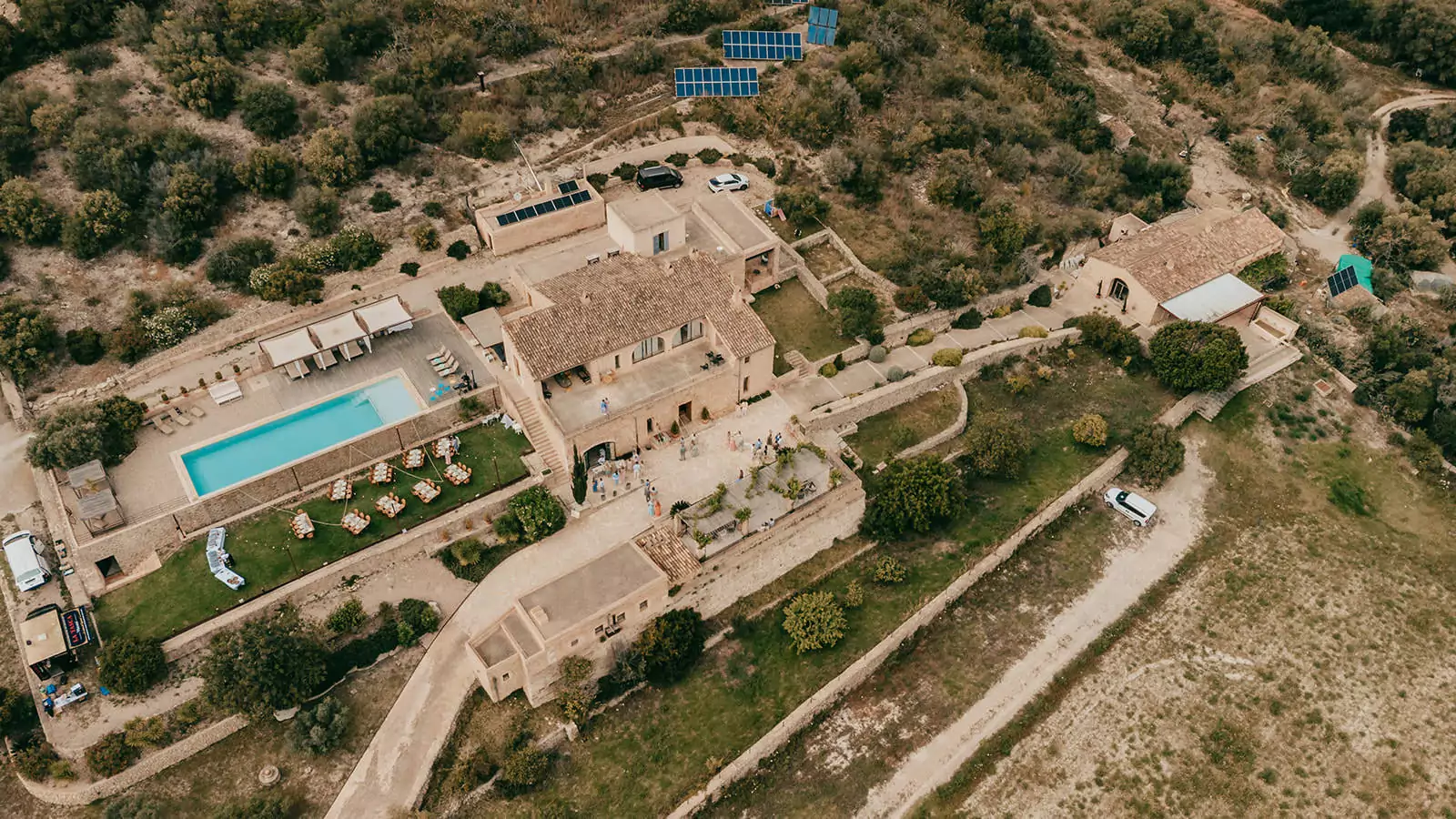 Luftaufnahme einer großen Steinvilla in Ses Voltes, Mallorca, perfekt für eine Hochzeit, mit Swimmingpool, üppigem Grün, Sonnenkollektoren auf einem nahe gelegenen Hügel und entlang der unbefestigten Straße geparkten Fahrzeugen.