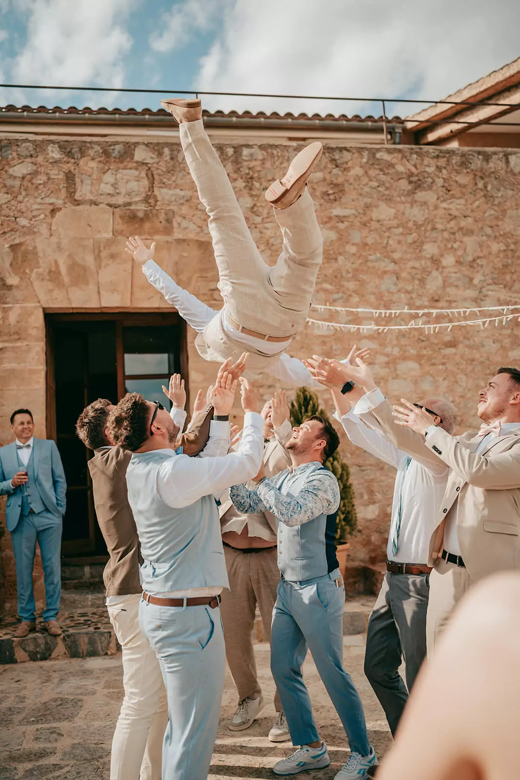Bei einer Ses-Voltes-Mallorca-Hochzeit wirft eine Gruppe von Männern in Anzügen einen anderen Mann in hellen Hosen und Schuhen bei einer Freiluftveranstaltung neben einem Steingebäude unter einem teilweise bewölkten Himmel freudig in die Luft.