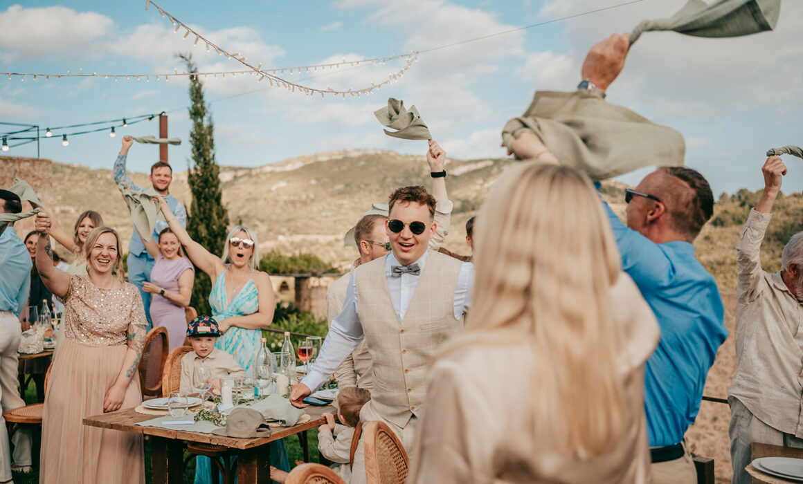Bei einer Ses-Voltes-Mallorca-Hochzeit winken die Gäste lächelnd und jubelnd mit Servietten in der Luft. Die festliche Szene zeigt geschmückte Tische unter einem blauen Himmel mit Blick auf die Berge, in deren Mittelpunkt ein Mann mit Sonnenbrille und beigem Anzug steht.