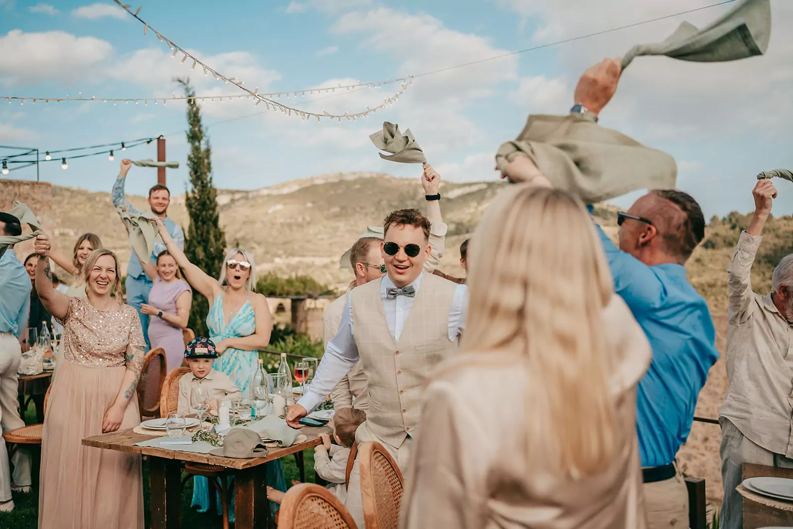 Bei einer Ses-Voltes-Mallorca-Hochzeit winken die Gäste lächelnd und jubelnd mit Servietten in der Luft. Die festliche Szene zeigt geschmückte Tische unter einem blauen Himmel mit Blick auf die Berge, in deren Mittelpunkt ein Mann mit Sonnenbrille und beigem Anzug steht.