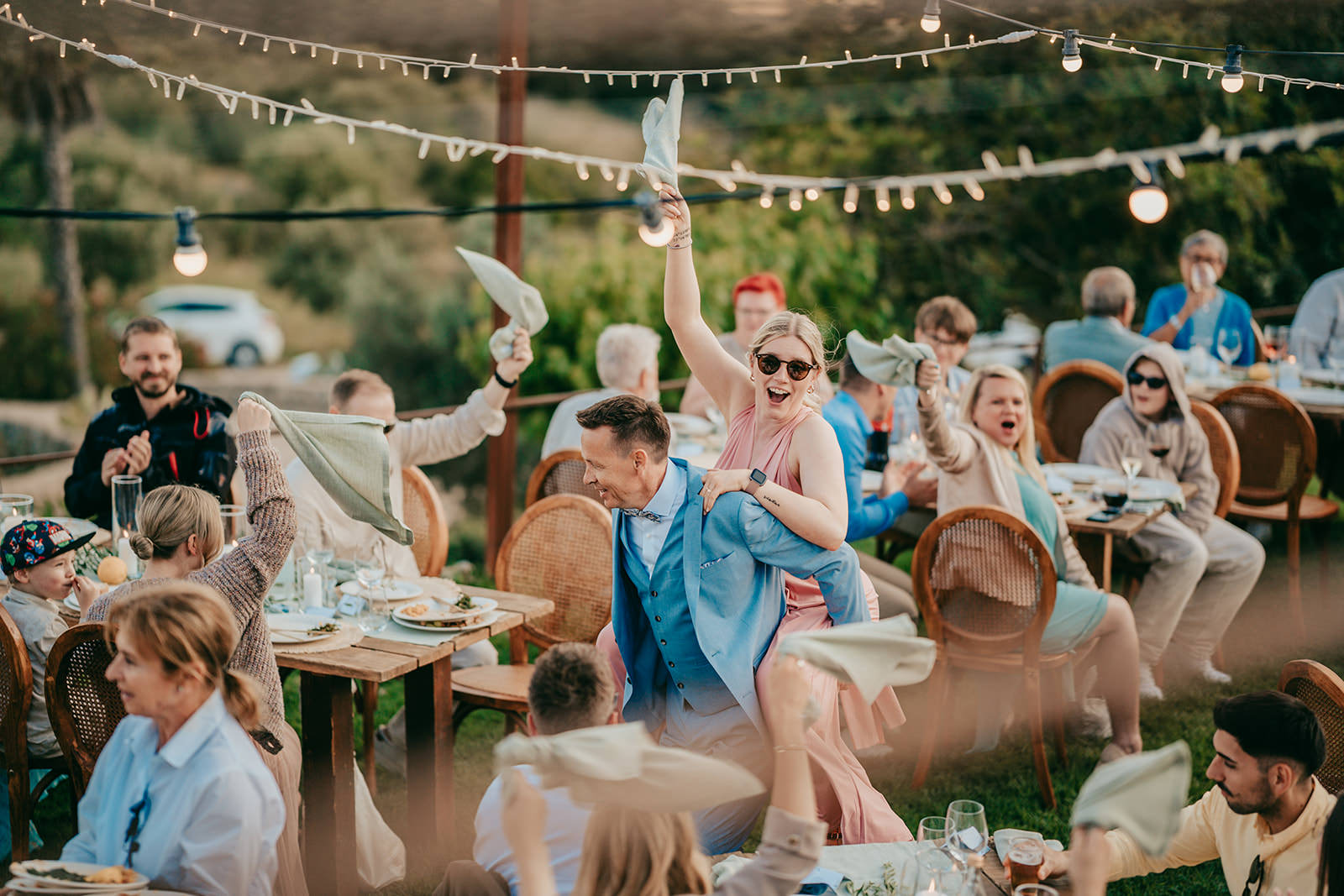 Eine fröhliche Hochzeitsfeier im Freien auf der Finca ses voltes mallorca, bei der die Gäste mit Servietten winken, während ein lächelndes Paar, das auf dem Rücken des anderen reitet, einen lebhaften Auftritt zwischen den mit Speisen und Getränken geschmückten Tischen unter Lichterketten hat.