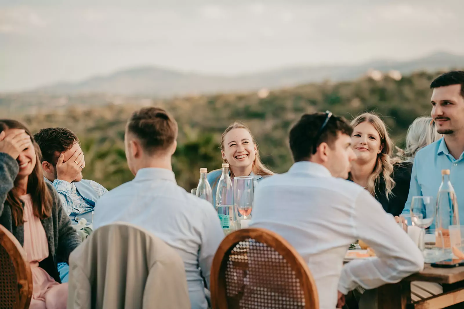 Eine Gruppe von Menschen sitzt um einen Tisch im Freien bei Ses Voltes Mallorca, lacht und genießt eine Mahlzeit mit Getränken. Im Hintergrund sind Berge und Grünflächen zu sehen, die eine entspannte, fröhliche Hochzeitsatmosphäre schaffen.