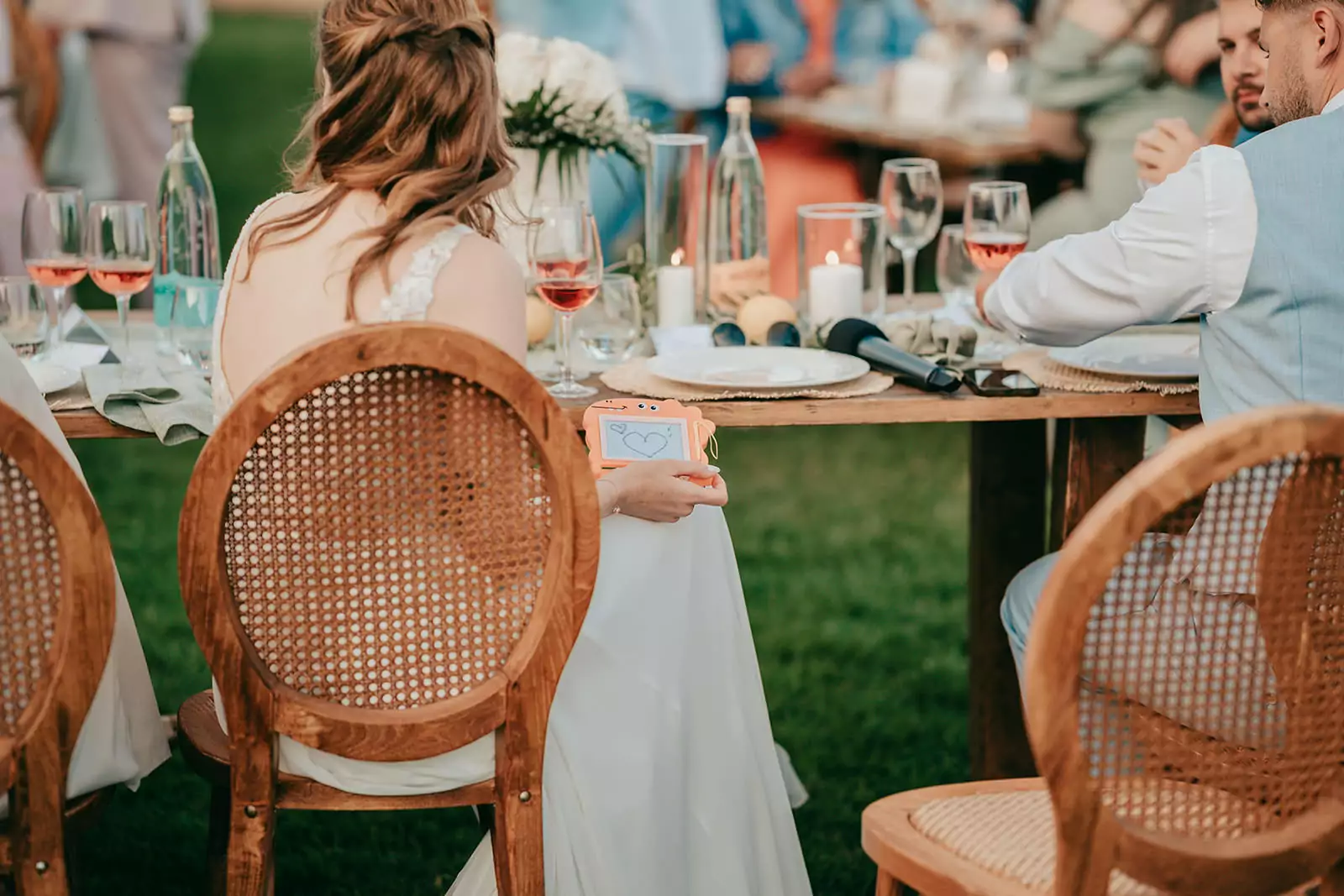 Eine Braut in einem weißen Kleid sitzt an einem Holztisch im Freien bei Ses Voltes Mallorca und hält eine kleine Karte mit einem blauen Herz in der Hand. Weingläser, Flaschen und Blumen schmücken die festliche Hochzeitsszene mit den in der Nähe sitzenden Gästen.