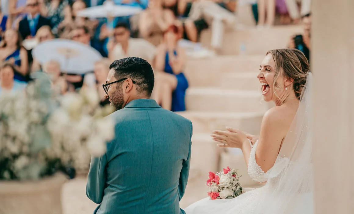 A bride in a white dress and veil sits beside a man in a blue suit, laughing and clapping during an outdoor wedding ceremony. The background shows guests seated on stone steps, watching the event.