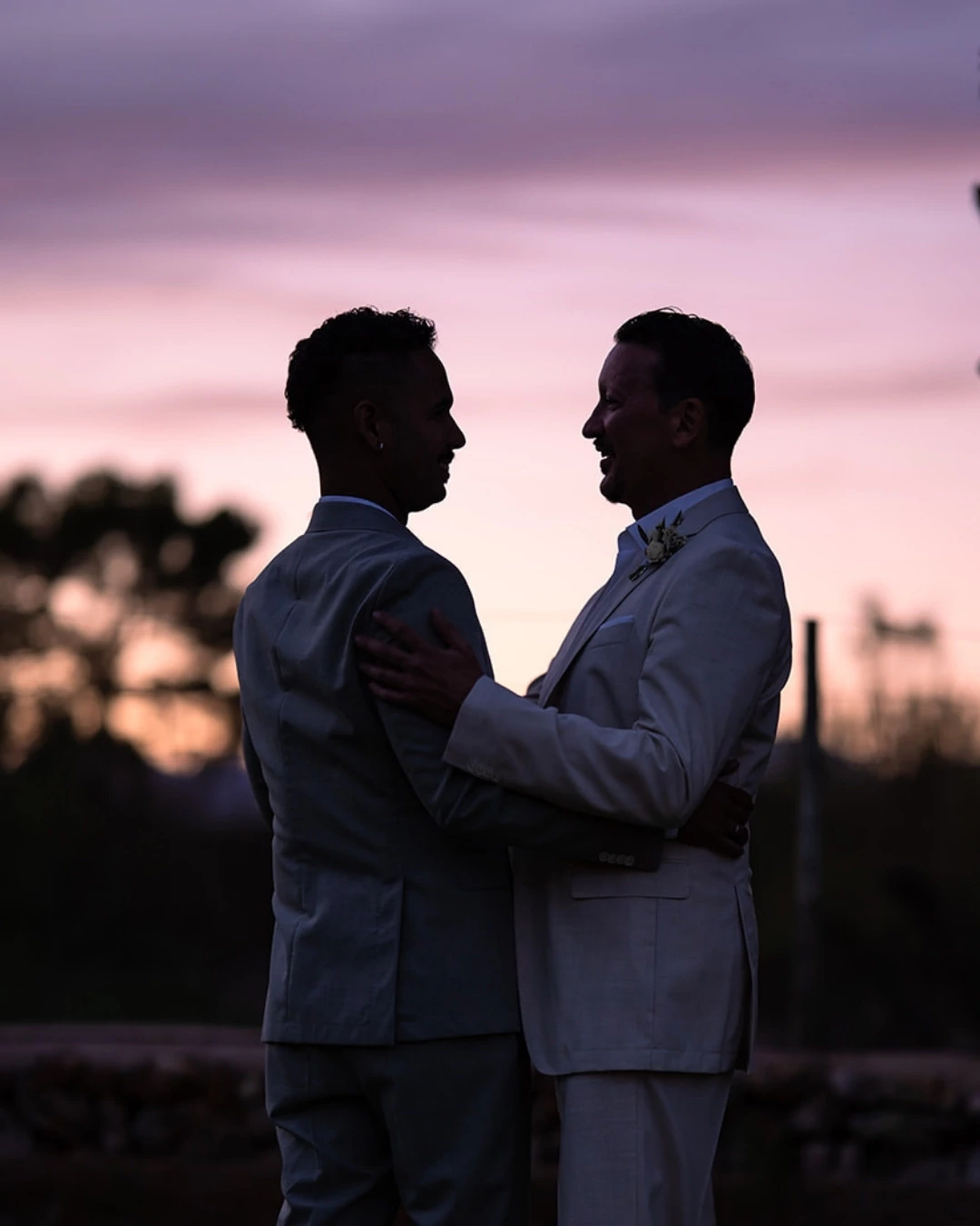 Zwei Männer in Anzügen umarmen sich bei Sonnenuntergang im Freien, vor einem lila-rosa Himmel - wunderschön eingefangen von einem Hochzeitsfotografen Mallorca auf der romantischen Finca Alaiar.