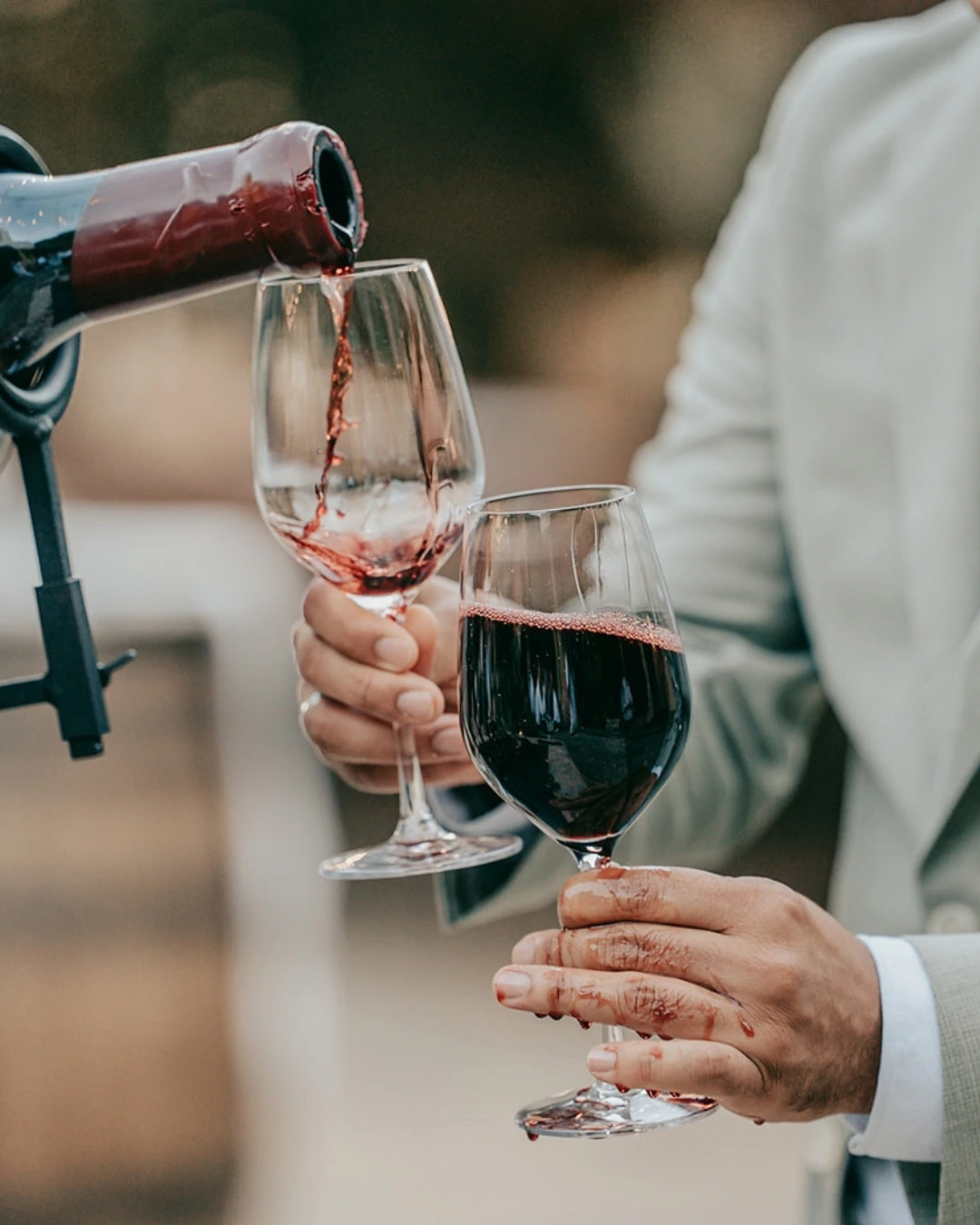 Eine Person in einem hellen Anzug, vielleicht ein Hochzeitsfotograf Mallorca, gießt Finca Alaiar Rotwein in ein Glas ein und hält ein zweites gefülltes Glas in der Hand. Der Wein ist auf ihre Hand und Finger verschüttet.