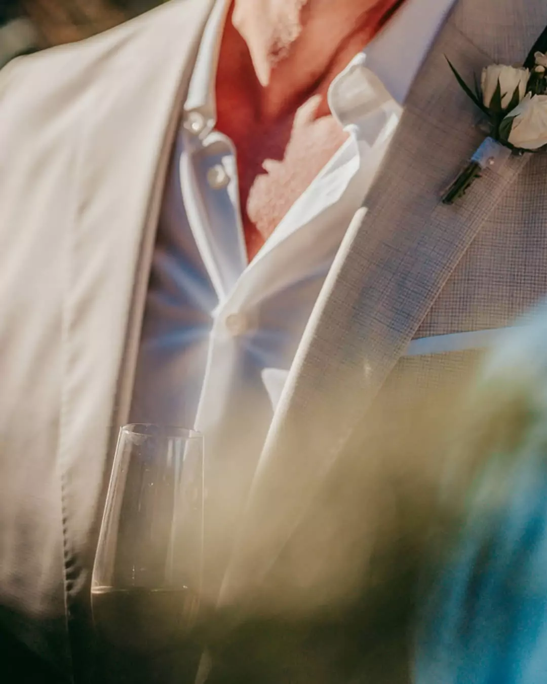 Eine Nahaufnahme einer Person in einem hellen Anzug mit weißem Hemd, Boutonnière und offenem Kragen, die ein Glas Champagner hält. Fotografiert von einem Hochzeitsfotograf Mallorca auf der Finca Alaiar, der Hintergrund ist sanft verschwommen.