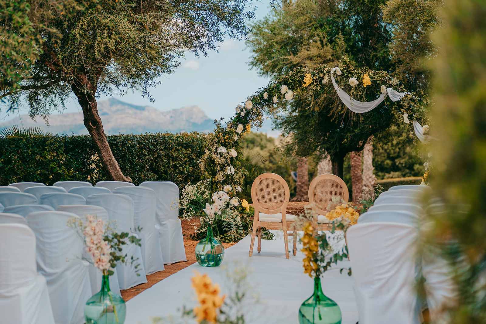 casal santa eulalia hochzeitslocation mallorca Hochzeit im Freien mit zwei Stühlen unter einem Blumenbogen, umgeben von Reihen weißgedeckter Stühle. Grüne Glasvasen mit gelben und weißen Blumen säumen den Gang. Im Hintergrund sind Bäume und Berge zu sehen.
