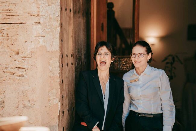 Zwei Frauen stehen lächelnd vor einer rustikalen Tür. Eine Frau, die einen dunklen Blazer trägt, lacht offen, während die andere, die ein gestreiftes Hemd mit einem Namensschild trägt, herzlich lächelt. Der Hintergrund ist ein sanft beleuchteter Innenraum.