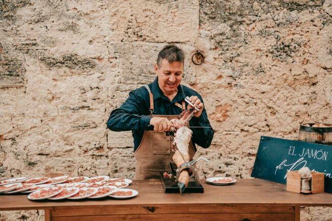 Ein Mann in einer Schürze schneidet auf einem Holztisch Schinken aus einer Keule, daneben stehen Teller mit Schinken. Er steht vor einer rustikalen Steinmauer, und rechts ist ein Schild mit der Aufschrift 