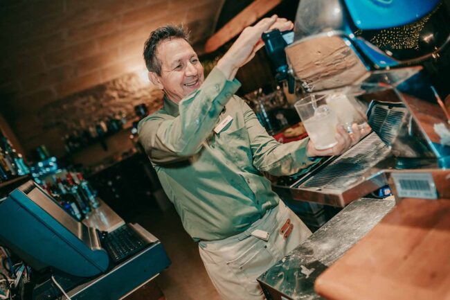 Ein lächelnder Barkeeper in grünem Hemd und beiger Schürze gießt ein Getränk aus einem Automaten in ein Glas hinter einer Bar ein, während im Hintergrund Flaschen und Barausstattung zu sehen sind.