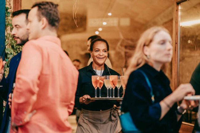 Ein lächelnder Kellner trägt ein Tablett mit rosafarbenen Getränken durch eine Menschenmenge bei einem geselligen Beisammensein oder einer Party. Die Szene ist lebhaft, die Gäste unterhalten sich und mischen sich in einem warm beleuchteten Raum.