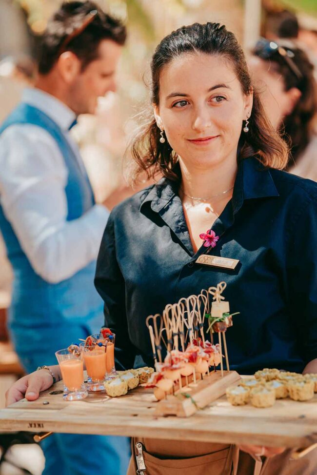 Eine Frau in einem marineblauen Hemd hält ein Holztablett mit verschiedenen Vorspeisen und kleinen Gläsern mit Suppe. Sie lächelt leicht, während die Menschen im Hintergrund verschwommen zu erkennen sind.
