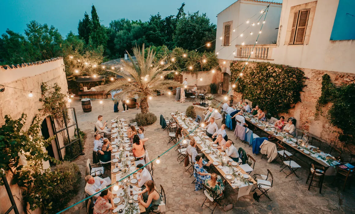 Ein Abendessen unter freiem Himmel auf der Finca Alaiar, bei dem die Gäste an langen Tischen unter Lichterketten in einem von Bäumen und rustikalen Gebäuden umgebenen Hof sitzen. Teller, Gläser und Dekorationen schmücken die Tische und schaffen eine magische Alaiar-Mallorca-Atmosphäre.