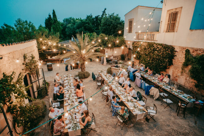 Ein Abendessen unter freiem Himmel auf der Finca Alaiar, bei dem die Gäste an langen Tischen unter Lichterketten in einem von Bäumen und rustikalen Gebäuden umgebenen Hof sitzen. Teller, Gläser und Dekorationen schmücken die Tische und schaffen eine magische Alaiar-Mallorca-Atmosphäre.