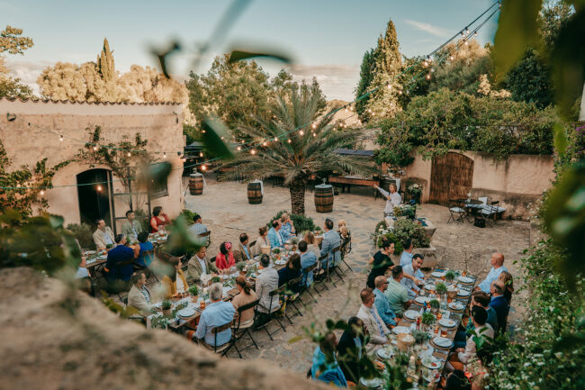 Eine Gruppe von Menschen genießt ein Treffen im Freien im rustikalen, mit Lichterketten geschmückten Hof der Finca Alaiar. Die Gäste sitzen an langen Tischen, die für ein Essen gedeckt sind, umgeben von Grün, Steinmauern und einer entspannten, festlichen Atmosphäre.