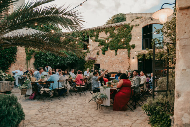 Eine Gruppe von Menschen speist gemeinsam an langen Tischen in einem rustikalen Innenhof der Finca Alaiar, der mit Lichterketten und Grünpflanzen geschmückt ist und eine festliche und entspannte Atmosphäre schafft.