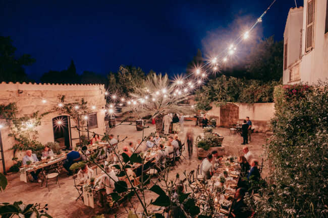 Eine abendliche Dinnerparty im Freien unter Lichterketten auf der Finca Alaiar. Die Gäste sitzen an Tischen in einem rustikalen Innenhof, umgeben von Bäumen, Pflanzen und alten Steingebäuden. Die warme, festliche Atmosphäre fängt den Charme von Alaiar Mallorca ein.