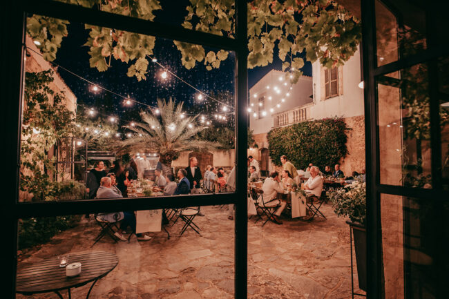 Auf der Finca Alaiar findet eine lebhafte Dinnerparty im Freien statt, bei der die Gäste unter Lichterketten in einem bezaubernden, von Grün und Steinmauern umgebenen Innenhof unter dem Nachthimmel Mallorcas speisen und sich unterhalten.