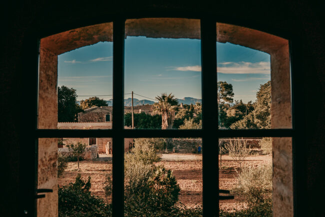Blick durch ein Fenster mit schwarzen Scheiben, das einen rustikalen Innenhof der Finca Alaiar, grüne Bäume und ferne Berge unter blauem Himmel zeigt. Der Fensterrahmen und die Steinmauer verleihen dieser Szene auf Alaiar Mallorca ihren Charme.
