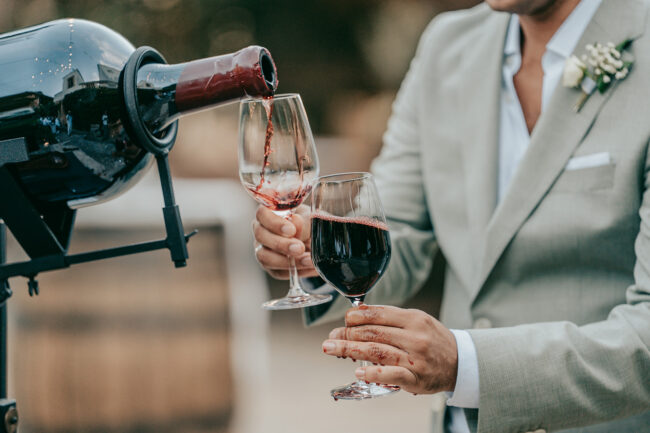 Eine Person in einem hellgrauen Anzug gießt Rotwein aus einer großen Flasche der Finca Alaiar in ein Glas, während sie ein weiteres Glas hält. Der sanft verschwommene Hintergrund deutet auf eine Alaiar-Mallorca-Außenanlage hin.