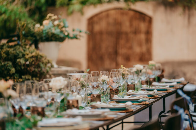 Ein elegant gedeckter Esstisch im Freien auf der Finca Alaiar ist mit Gläsern, Tellern, Servietten und Blumensträußen mit Grünzeug bestückt und steht vor einer rustikalen Holztür, umgeben von üppigem Gartenlaub.