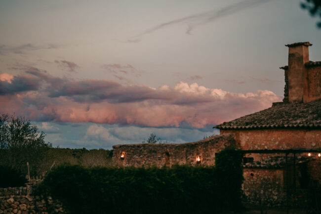 Ein rustikales Steinhaus mit Ziegeldach auf der Finca Alaiar erstrahlt bei Sonnenuntergang in warmem Licht. Dahinter ziehen rosa und lila Wolken über den Abendhimmel und schaffen eine ruhige Atmosphäre, die perfekt für eine Alaiar Mallorca Hochzeit ist.