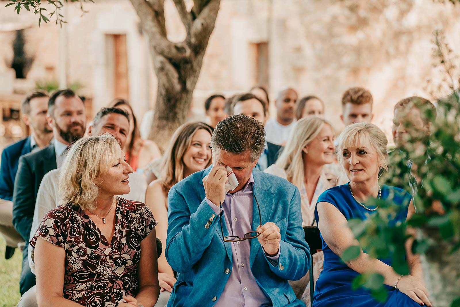 mallorca hochzeit alaiar 300 websize Eine Gruppe von Menschen sitzt im Freien, halb formell gekleidet, lächelt und unterhält sich. Ein Mann im Vordergrund, der eine blaue Jacke trägt, wischt sich über die Augen und hält eine Brille. Im Hintergrund sind Bäume und ein verschwommenes Gebäude zu sehen.