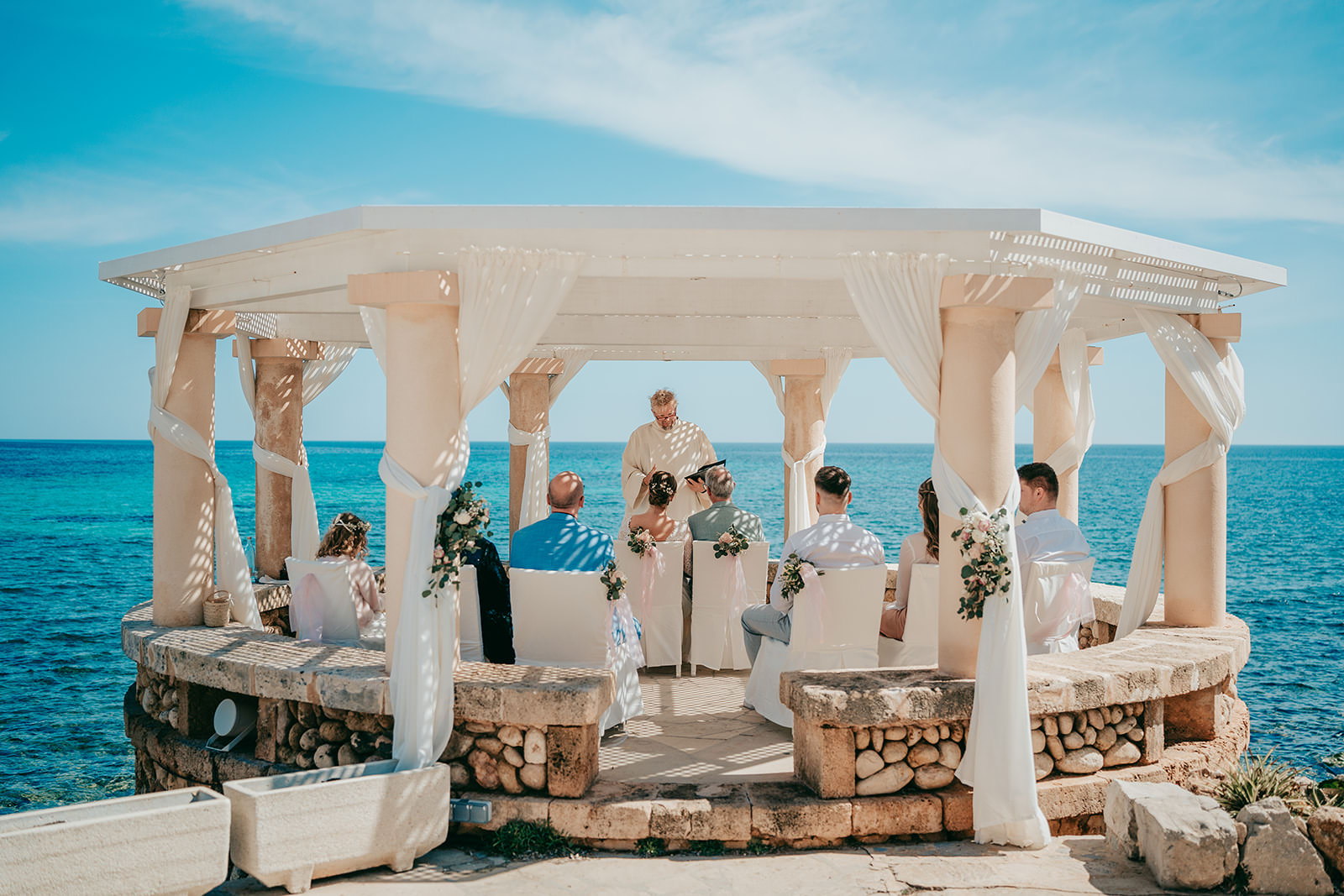 Eine kleine Hochzeitszeremonie findet unter einem weißen Pavillon am Meer in Port verd del mar statt, wobei die Gäste sitzen und der Trauzeuge vor dem Paar steht. Der Himmel ist klar und das Meer leuchtet hellblau im Hintergrund.