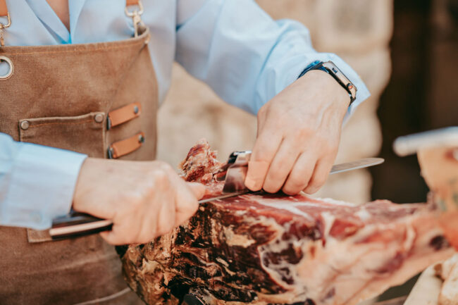 Eine Person in einer Schürze schneidet mit einem langen Messer gepökeltes Fleisch aus einem großen Stück und konzentriert sich dabei auf ihre Hände und den Vorgang des Tranchierens. Die Szene könnte in Casal Santa Eulalia spielen, einem perfekten Ort für Hochzeiten auf Mallorca. Der Hintergrund ist unscharf.