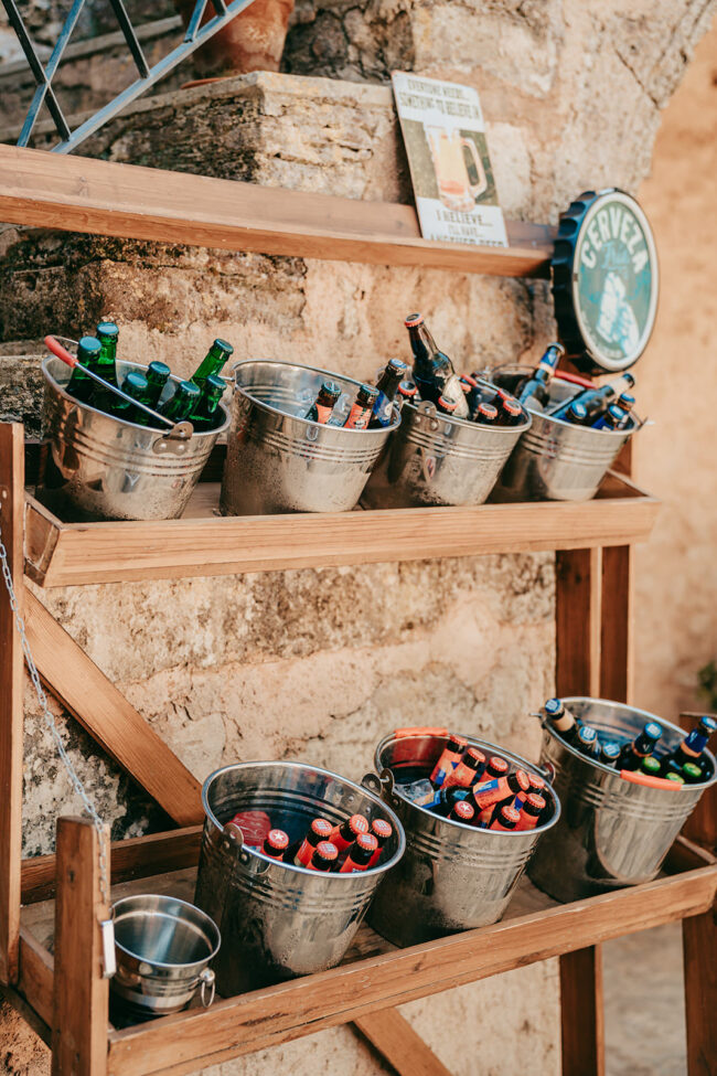 Metalleimer, gefüllt mit verschiedenen Getränken in Flaschen, hauptsächlich Bier, sind auf Holzregalen vor einer rustikalen Steinmauer in Casal Santa Eulalia angeordnet. So entsteht eine zwanglose Getränkestation im Freien, die sich perfekt für eine Hochzeit auf Mallorca eignet.