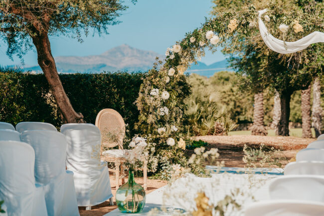 Hochzeit im Freien mit weißen Stühlen, einem Blumenbogen und Blick auf die Berge in Casal Santa Eulalia. Das Sonnenlicht und die grüne Vegetation unterstreichen diese hochzeit auf Mallorca Location und schaffen eine friedliche, malerische Szene.