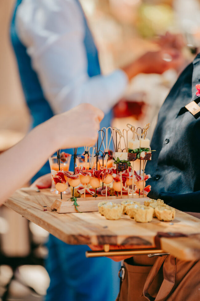 Bei einer Veranstaltung in Casal Santa Eulalia hält eine Person ein Holztablett mit verschiedenen Häppchen auf Spießen und kleinem Gebäck, während eine andere Hand einen Spieß aufnimmt - perfekt für eine Hochzeit auf Mallorca inmitten elegant gekleideter Gäste.