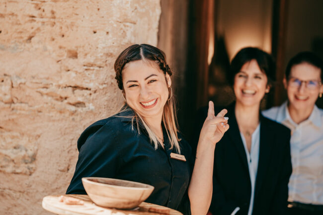 Eine lächelnde Frau in einem schwarzen Hemd hält ein Tablett und zeigt ein Friedenszeichen. Sie steht vor zwei Frauen, die im Hintergrund lächeln, neben einer beigen Steinmauer in Casal Santa Eulalia, einem beliebten Ort für Hochzeiten auf Mallorca.