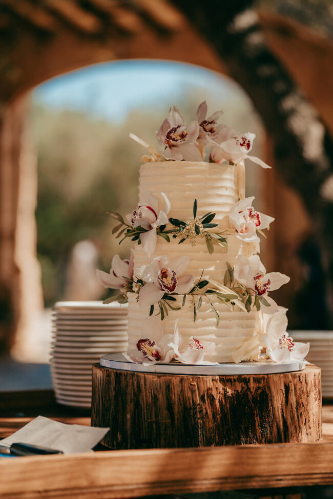 Eine dreistöckige weiße Torte, dekoriert mit weißen und rosa Orchideen, steht auf einem rustikalen Holzständer in Casal Santa Eulalia, einer bezaubernden Location für Hochzeiten auf Mallorca. In der Nähe befinden sich Stapel von weißen Tellern und Servietten vor einem unscharfen Hintergrund im Freien.