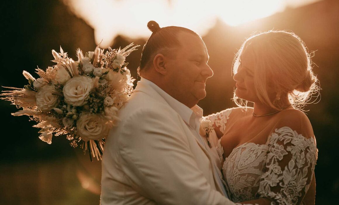 Ein Paar in Hochzeitskleidung umarmt sich bei Sonnenuntergang im Freien. Der Bräutigam trägt einen weißen Anzug und die Braut in einem Spitzenkleid hält einen Strauß weißer Blumen. Warmes Sonnenlicht beleuchtet sie sanft von hinten.