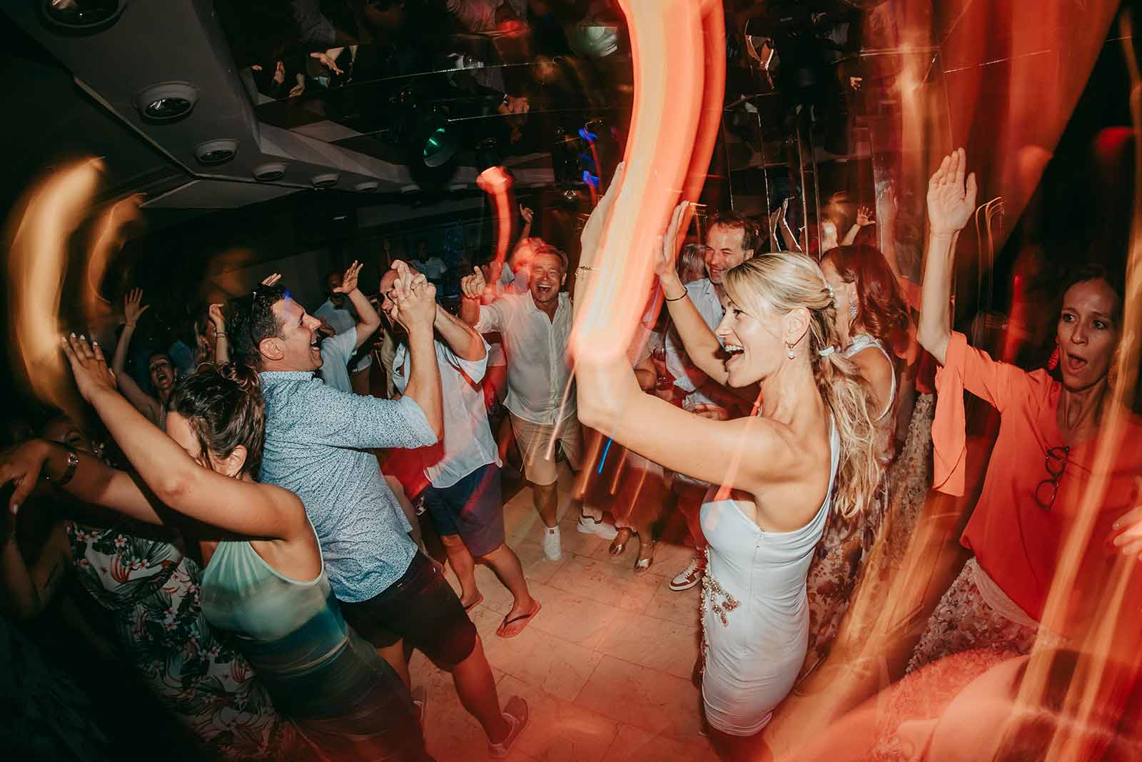 hochzeit mallorca zeitplan party Eine lebhafte Gruppe von Menschen tanzt und feiert auf einer Party, wobei bunte Bewegungsunschärfen und Lichter eine dynamische, energiegeladene Atmosphäre schaffen. Einige Personen haben die Arme erhoben und lächeln freudig.