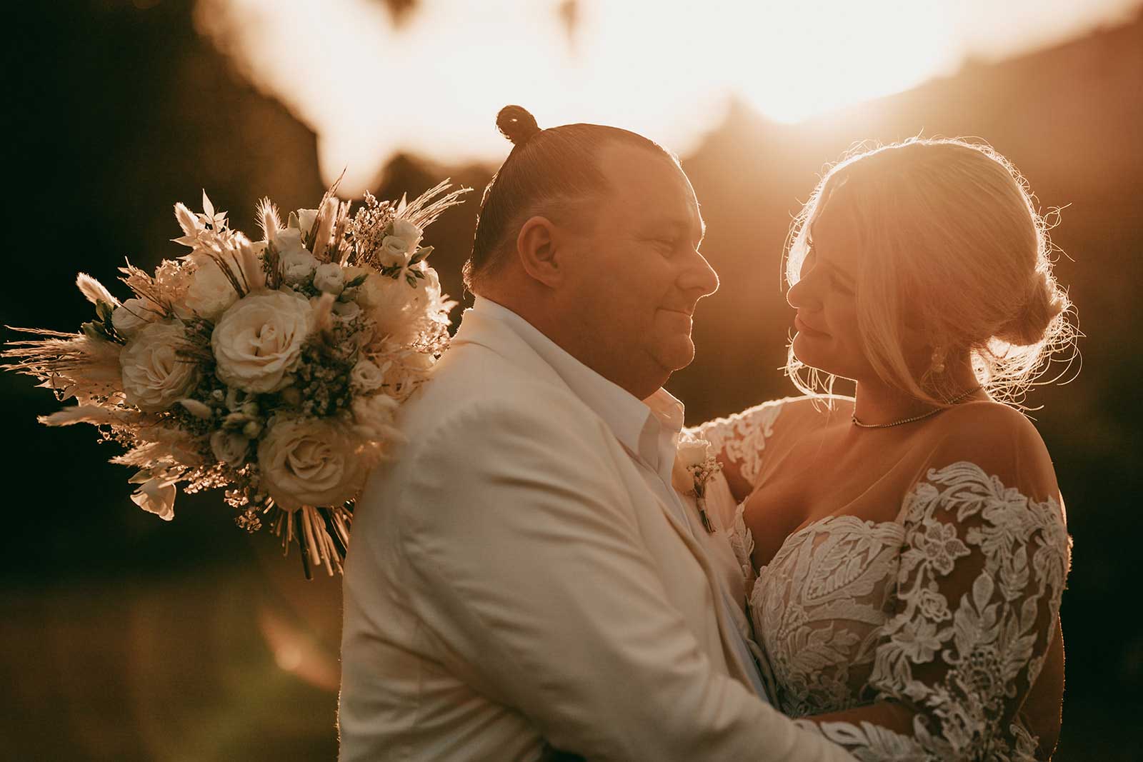hochzeit mallorca zeitplan Ein Paar in Hochzeitskleidung umarmt sich bei Sonnenuntergang im Freien. Der Bräutigam trägt einen weißen Anzug und die Braut in einem Spitzenkleid hält einen Strauß weißer Blumen. Warmes Sonnenlicht beleuchtet sie sanft von hinten.